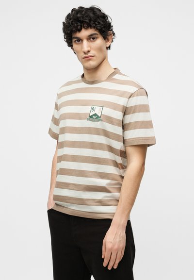 Νέος άνδρας με σγουρά μαλλιά, φορώντας ριγέ μπλουζάκι σε μπεζ και λευκό με ένα μικρό σήμα στο στήθος και μαύρο παντελόνι, στέκεται με τα χέρια μερικώς στις τσέπες.