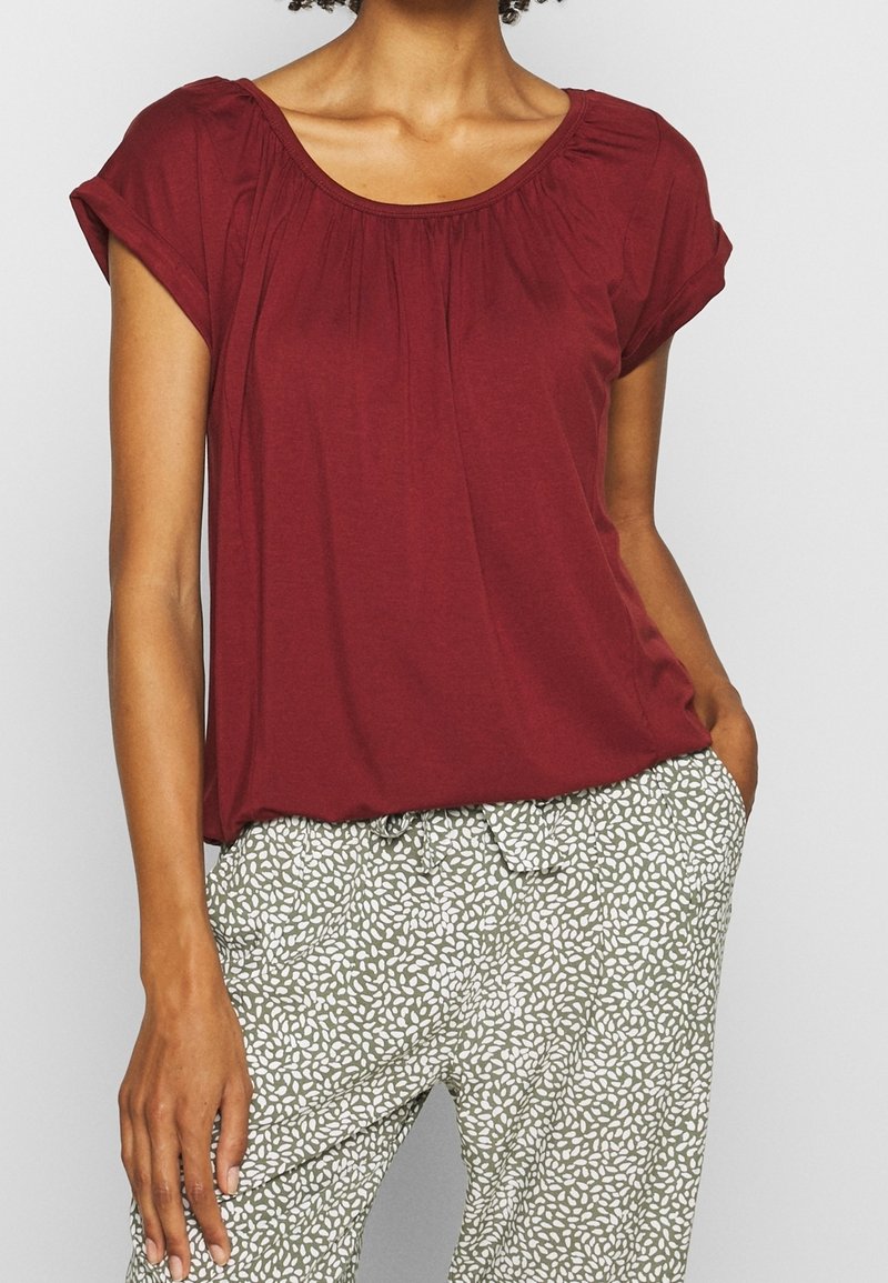 Burgunderfarbenes kurzärmliges Oberteil mit gerafftem Kragen, kombiniert mit hellgrünen Hosen mit einem weißen Blattmuster. Sanfter, leichter Stoff.
