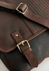 Ruskea nahkainen postimieslaukku, jossa on teksturoitu läppä ja messinkiset metalliosat. Mukana solki ja ommellut yksityiskohdat kestävyyttä varten.