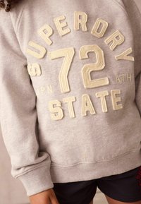 Šedá mikina so zvýrazneným krémovým nápisom "SUPERDRY 72 STATE" a rebrovanými manžetami. Nosená cez tmavé šortky, čo vytvára uvoľnený vzhľad.