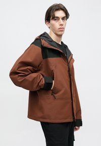 Καφέ και μαύρο δίχρωμο μπουφάν με φερμουάρ στο μπροστινό μέρος, ρυθμιζόμενες μανσέτες και τσέπες. Το υλικό φαίνεται υφή και ανθεκτικό στις καιρικές συνθήκες.