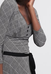 Svart och vit rutig klänning med långa ärmar, knapppdetaljer och snodd midja. Tyget verkar ha en struktur med en figurnära silhuett.