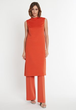 Mouwloze oranje jurk met een gestructureerd, gewatteerd patroon en een hoge halslijn. Gecombineerd met een bijpassende wijde broek.