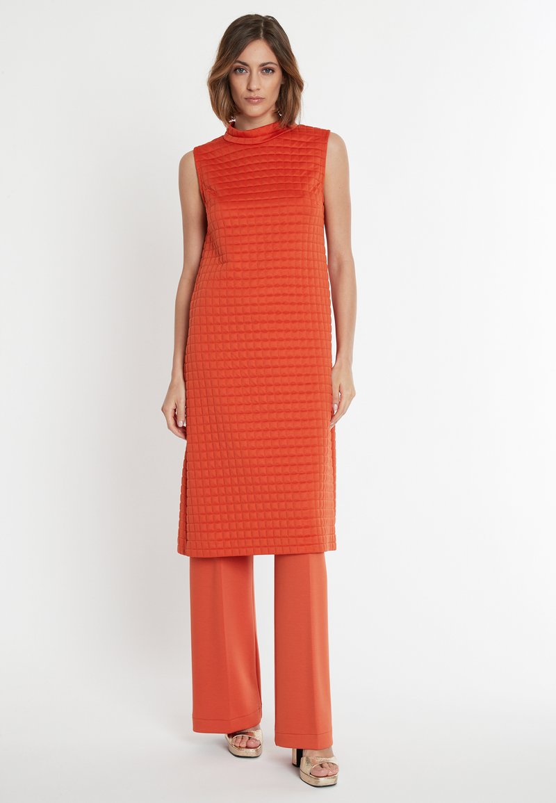 Ärmelloses oranges Kleid mit einem strukturierten, gesteppten Muster und hohem Kragen. Kombiniert mit passenden, weit geschnittenen Hosen.