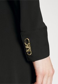 Robe noire avec une texture lisse, ornée d'un accent en métal doré affichant les initiales de la marque au niveau du poignet.