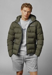 Mladic s kovrčavom kosom nosi maslinasto zeleni puffer jaknu preko sive Red Bull Racing dukserice i odgovarajućih kratkih hlača.