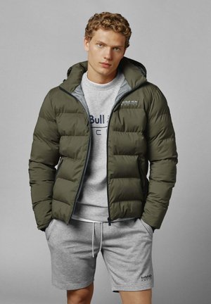 Jonge man met krullend haar die een olijfgroen pufferjack over een grijze Red Bull Racing sweatshirt en bijpassende shorts draagt.