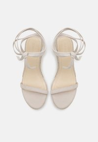 Beige semskede sandaler med flate såler, med tynne stropper over tærne og rundt ankelen, knyttet i en sløyfe. Myk tekstur, minimalistisk design.