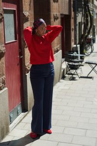 Rød langærmet sweater, marineblå vidde bukser, røde flade sko og et mønstret hovedtørklæde. Sat op imod en stenmur og dør.