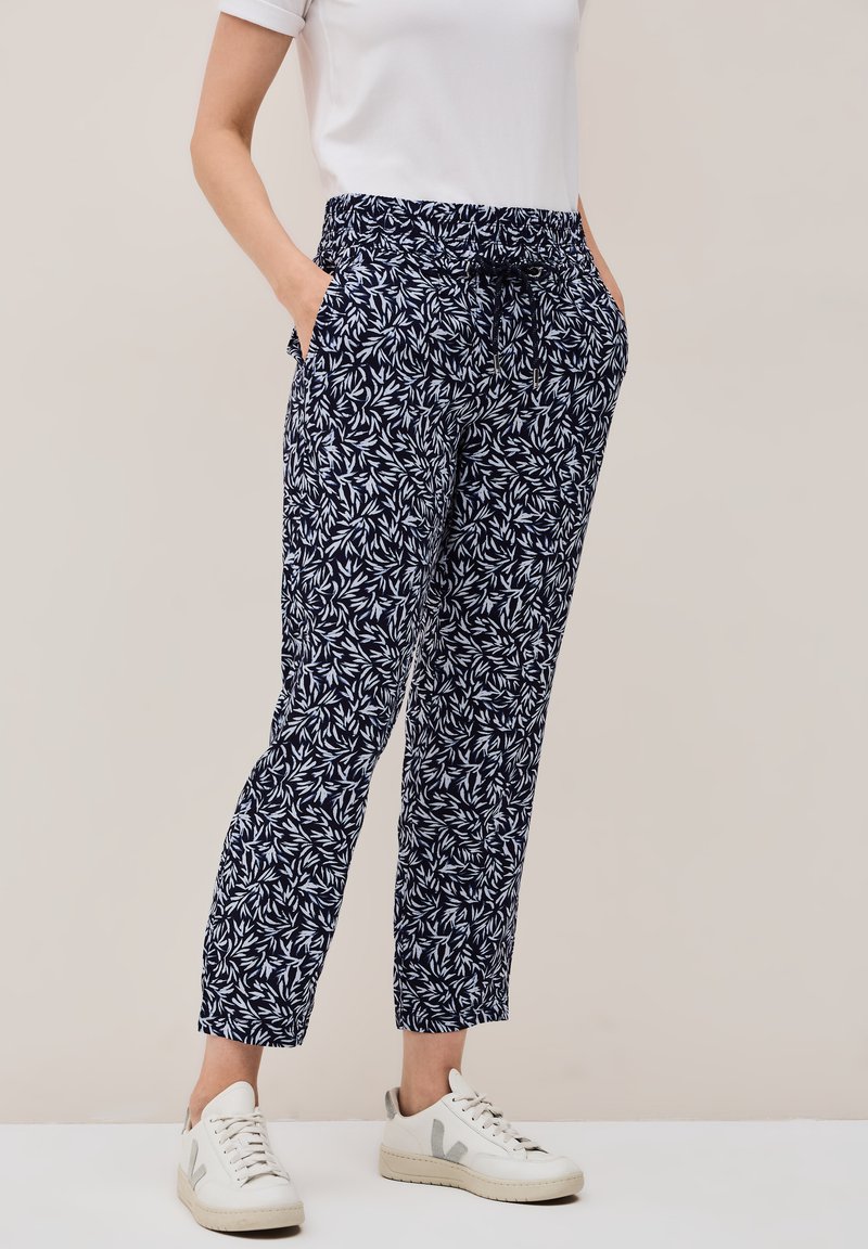 Pantaloni navy con un motivo foglioso bianco, cintura elastica e tasche laterali, abbinati a sneakers bianche. Tessuto leggero, vestibilità comoda.