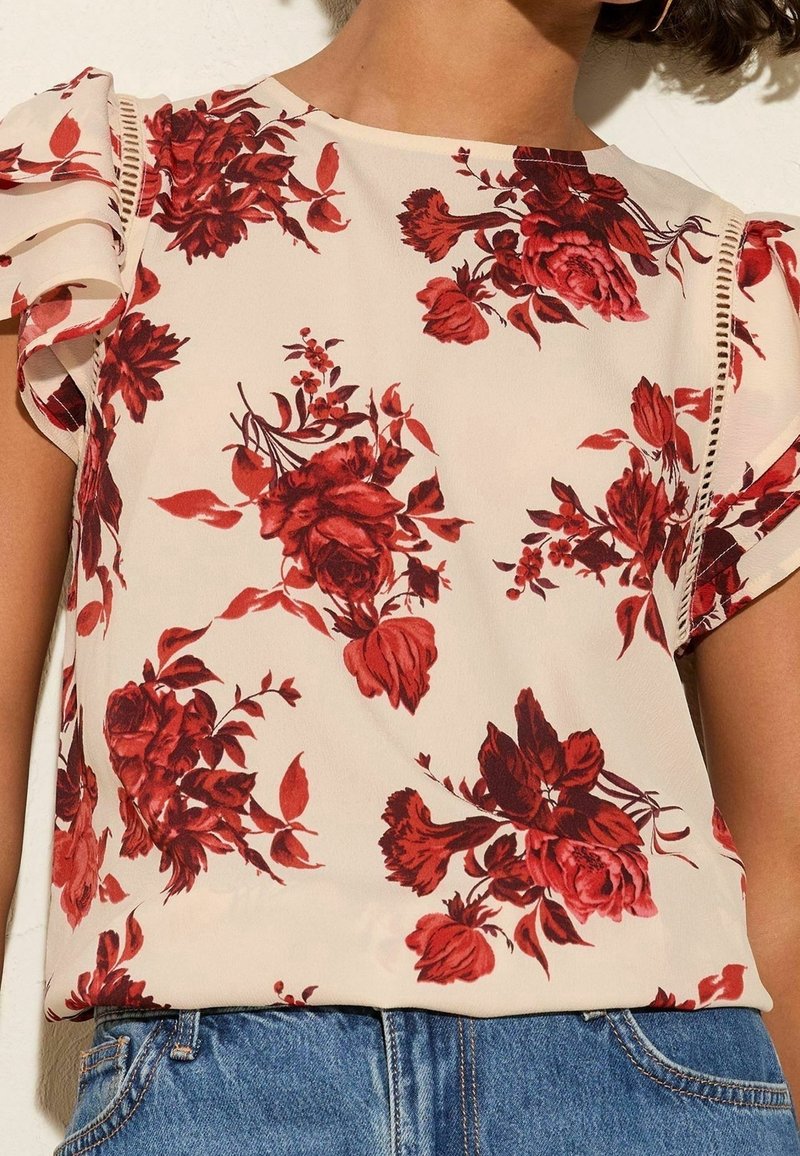Femme portant un chemisier crème avec imprimé floral rouge et manches courtes à volants, associé à un jean en denim bleu.