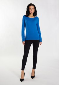 Kék kötött pulóver hosszú ujjal és lekerekített nyakkivágással, fekete slim-fit nadrággal és fekete hegyes orrú magassarkúval párosítva, világos háttér előtt.