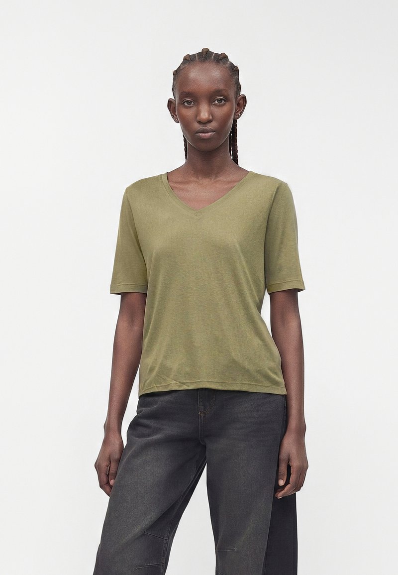 Frau mit geflochtenem Haar, die ein olivgrünes kurzärmeliges V-Neck-Shirt und dunkelgraue Hosen trägt, steht vor einem schlichten hellen Hintergrund.