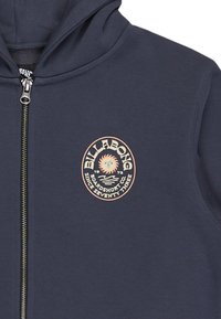 Felpa con zip di colore blu navy realizzata in tessuto morbido, con un logo circolare Billabong sul lato sinistro del petto e un design solare in tonalità chiare.