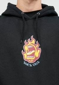 Μαύρη φόρμα με κουκούλα και κορδόνια με μεταλλικές άκρες, που διαθέτει μια πολύχρωμη γραφική απεικόνιση ενός στόματος περιτριγυρισμένου από φλόγες και το κείμενο "SANTA CRUZ" από κάτω.