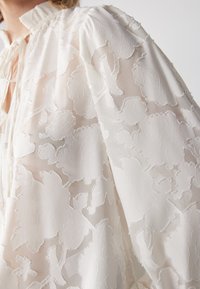 Blusa blanca con bordado floral, tela ligera, cuello con volantes y detalle de lazo. Presenta mangas abullonadas con acentos texturizados.