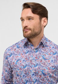 Langsleeve shirt met een levendig paisleypatroon in rood, blauw en paars op een witte achtergrond, gemaakt van lichte katoen.