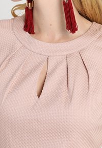 Donna che indossa un top rosa chiaro strutturato con scollatura a chiave e lunghe orecchini a nappina rossi.