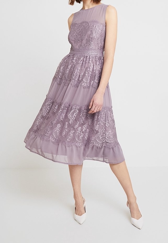 En lila klänning med lager av spets och en figursydd bodice med hög halsringning. Kjolen är flödande med A-linjeform och delikata spetsdetaljer.