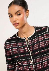 Černá bunda s červeno-bílým kostkovaným vzorem, texturovaná tkanina, kulatý výstřih, dekorativní krémová lemování a kulaté přední knoflíky.