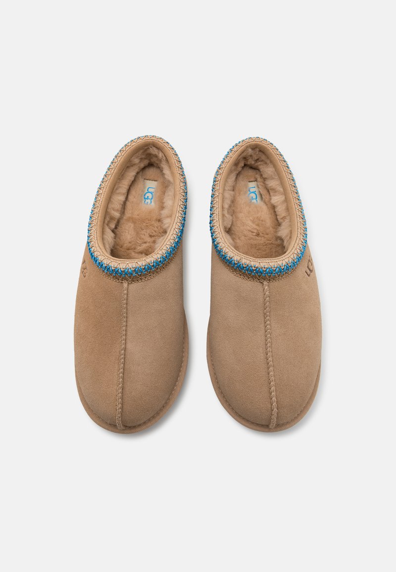 Bruine suède pantoffels met een zachte beige bontvoering, voorzien van een decoratieve blauwe geborduurde rand rond de kraag en eenvoudige stikdetails.