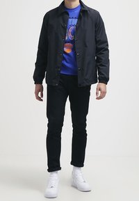 Giacca leggera nera su una felpa grafica blu, abbinata a jeans neri e sneakers bianche. Design semplice con chiusura a bottoni a scatto.