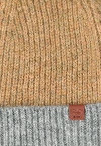 Czapka dziergana z tanowym wierzchem i szarym dołem w prążki, z brązową skórzaną metką z tłoczonym napisem. Miękka tekstura i ciepły design.