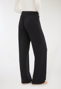 Donna che indossa pantaloni neri a vita alta e gamba larga e una maglietta bianca a maniche lunghe, in piedi a piedi nudi su uno sfondo chiaro.