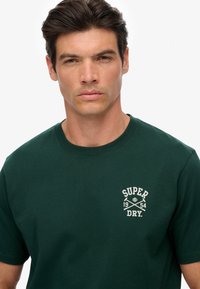 Donkergroene katoenen t-shirt met ronde hals en een klein wit grafisch ontwerp op de linkerborst met de tekst "SUPER DRY" en gekruiste bijlen.