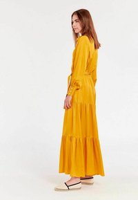 Longue robe jaune avec des manches longues, design à volants, poignets boutonnés et ceinture à la taille. Associée à des espadrilles beiges.