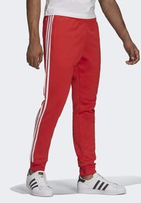 Pantaloni da tuta rossi con vita elastica, tasche laterali e tre strisce bianche lungo le cuciture esterne. Abbinati a sneakers bianche con accenti neri.