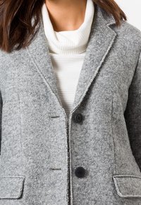 Szary blezer z mieszanki wełny o fakturowanej powierzchni, z klapami z wcięciem, dwoma czarnymi guzikami i dwoma bocznymi kieszeniami. Nosi się go na białym prążkowanym golfie.