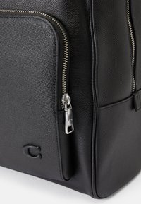Mochila de couro preta com acabamento texturizado, apresentando um bolso com fecho de correr proeminente, ferragens brilhantes e um logotipo em relevo discreto.