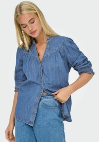 Camicia in denim con colletto arricciato e maniche con risvolto. Colore azzurro chiaro e vestibilità rilassata, con dettagli di cucitura lungo le cuciture.