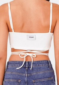 Top corto bianco a coste con spalline regolabili, dotato di una chiusura sul retro con nastro e una patch con logo vicino alla vita. Pantaloncini di jeans visibili sotto.