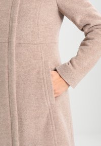 Cappotto in lana beige con una morbida texture, caratterizzato da un taglio dritto, due tasche laterali e dettagli di cucitura lungo il davanti e le maniche.