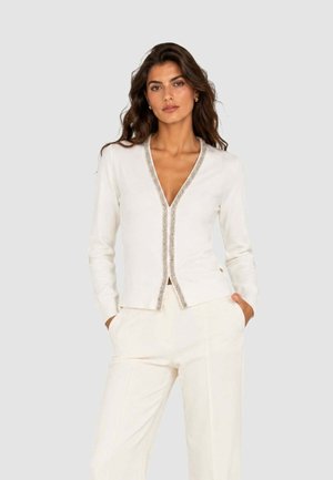 Donna con lunghi capelli scuri che indossa una giacca bianca con scollo a V con rifiniture argentate e pantaloni bianchi abbinati, mani nelle tasche, sfondo neutro.