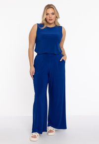 Blå ærmeløs jumpsuit med en flydende top og brede ben. Blødt stof, der har sidelommer. Bæres med hvide korsrem sandaler.