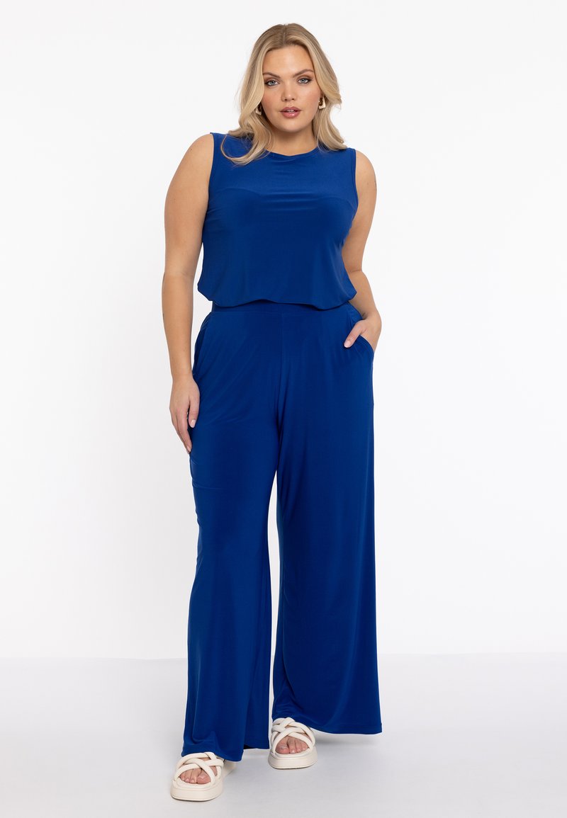 Blå ærmeløs jumpsuit med en flydende top og brede ben. Blødt stof, der har sidelommer. Bæres med hvide korsrem sandaler.