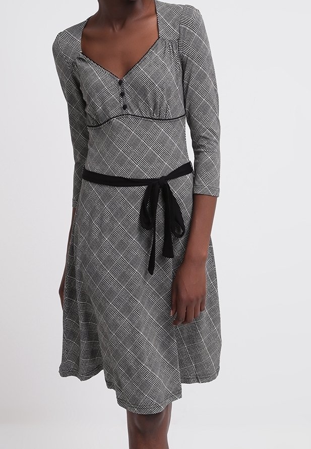 Svart och vit rutigt klänning med trekvartsärmar, V-halsringning, knappar framtill och ett svart bälte som framhäver midjan. Mjuk tygtextur.