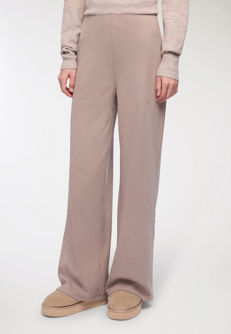 Szerokie, wysokie spodnie w kolorze miękkiego beżu, z prążkowaną teksturą i subtelnym wzorem w kratę, połączone z tanimi butami wsuwanymi.