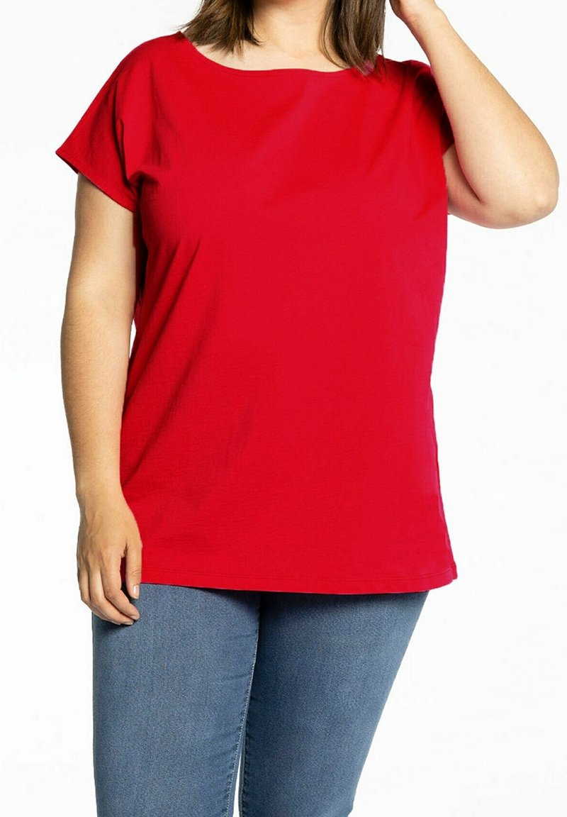 Femme portant une chemise rouge à manches courtes et un jean bleu, debout devant un fond blanc uni.
