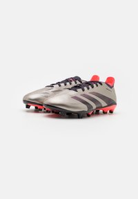 Scarpe da calcio con un upper argentato metallico, strisce nere e viola, accenti rossi sul tallone e una superficie texturizzata per la presa sull'erba.