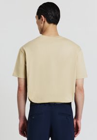 Uomo con capelli corti e scuri, che indossa una maglietta beige liscia e pantaloni blu scuro, mostrato di spalle su uno sfondo neutro.