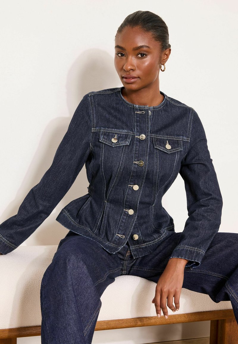 Žena sediaca na lavičke, oblečená v tmavomodrej rifľovej bunde a džínsoch, hľadí do kamery s neutrálnym výrazom v jednoduchom vnútornom prostredí.