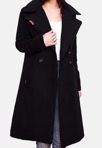 Donna che indossa un lungo cappotto nero doppiopetto con ampi risvolti sopra una maglietta bianca e jeans blu, tenendo il colletto del cappotto con una mano.
