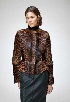 Femme aux cheveux bruns courts portant une veste en fausse fourrure à motif léopard sur une jupe en cuir noire sur un fond uni.