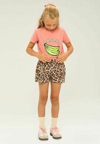 Rožinė marškinė su žalia banano grafika ir "BANANA" tekstu, derinamos su smėlio spalvos leopardų rašto šortais. Šviesiai rožinės sportbačiai ir kojinės užbaigia aprangą.