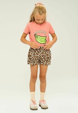 Camiseta rosa con gráfico de plátano verde y texto "BANANA", combinada con pantalones cortos de leopardo beige. Zapatillas rosa claro y calcetas completan el atuendo.