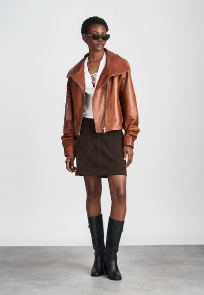 Brown leather jacket with a wide collar, white textured top, brown suede mini skirt, and black knee-high boots. Dark sunglasses complete the look.